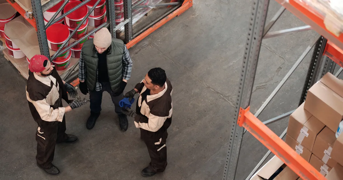 Vista desde arriba de tres trabajadores con overol y guantes conversando de pie en el pasillo de un almacén, entre racks con cubetas y cajas