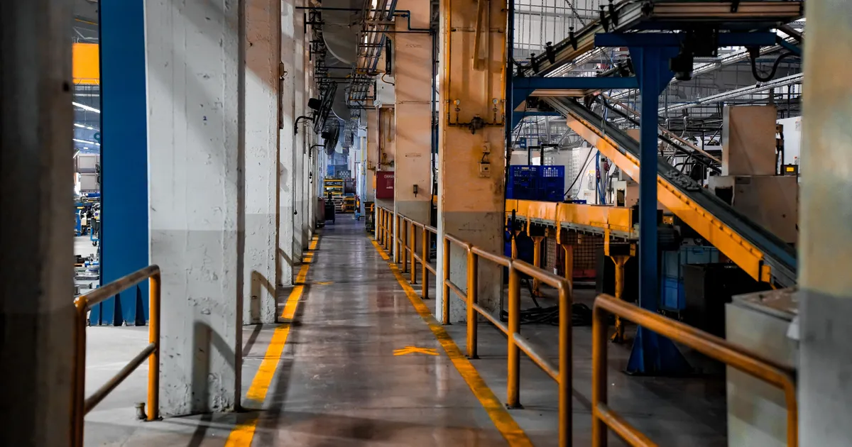 Pasillo peatonal vacío con barandal amarillo y banda transportadora al fondo, en el interior de una nave industrial sin personal en el turno