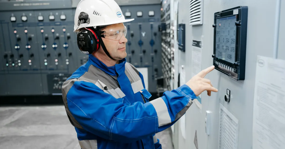 Técnico con casco blanco, lentes de seguridad y overol azul opera un tablero eléctrico con pantalla digital en una sala de control industrial