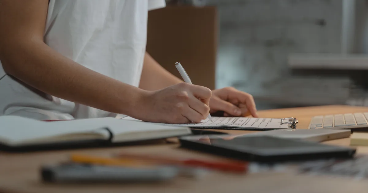 Manos llenan con pluma un formato en tabla sujetapapeles sobre un escritorio con libreta y teclado