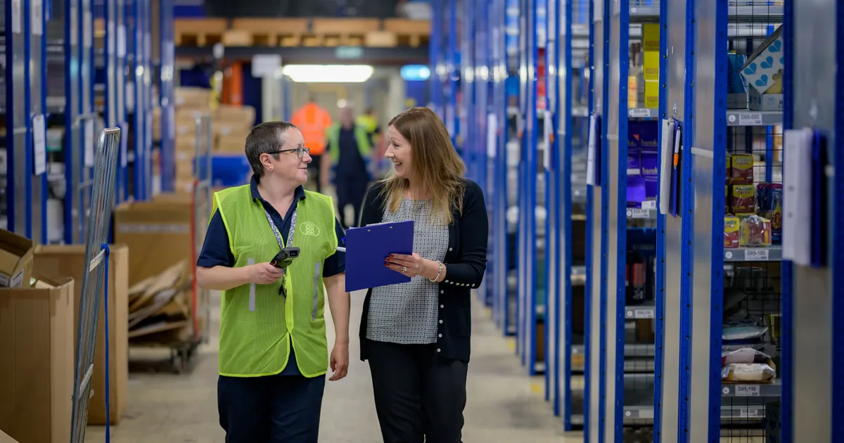 Dos trabajadoras conversan en el pasillo de un almacén: una con chaleco reflectante y escáner, la otra con una tabla portapapeles azul