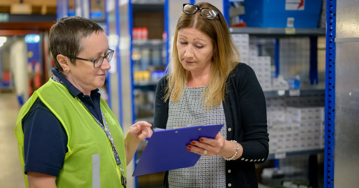 Dos trabajadoras de un almacén industrial revisan juntas una hoja sujeta a una tabla, de pie junto a los racks metálicos