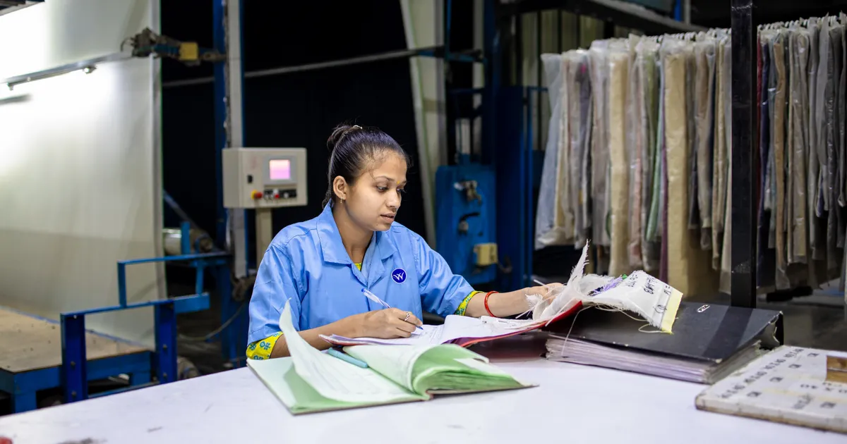 Trabajadora con uniforme azul escribe en un formato sobre una mesa dentro de una nave industrial, rodeada de carpetas y expedientes apilados