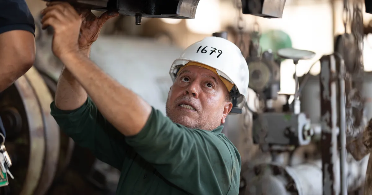 Técnico de mantenimiento con casco blanco numerado y camisola verde revisa desde abajo una pieza de maquinaria pesada dentro de un taller