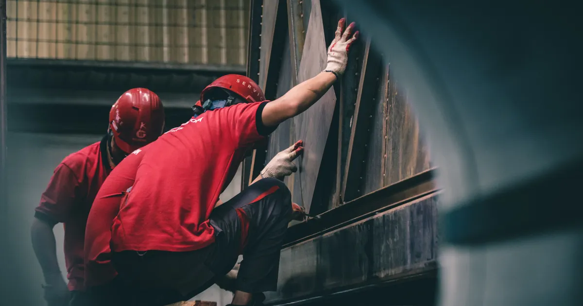 Dos operarios con casco rojo y guantes de trabajo sujetan una placa metálica contra una estructura de acero dentro de un taller industrial