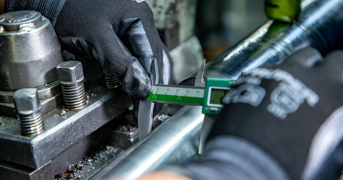 Manos con guantes negros miden una barra metálica con un vernier digital verde sobre la mesa de una máquina, entre virutas y tornillería