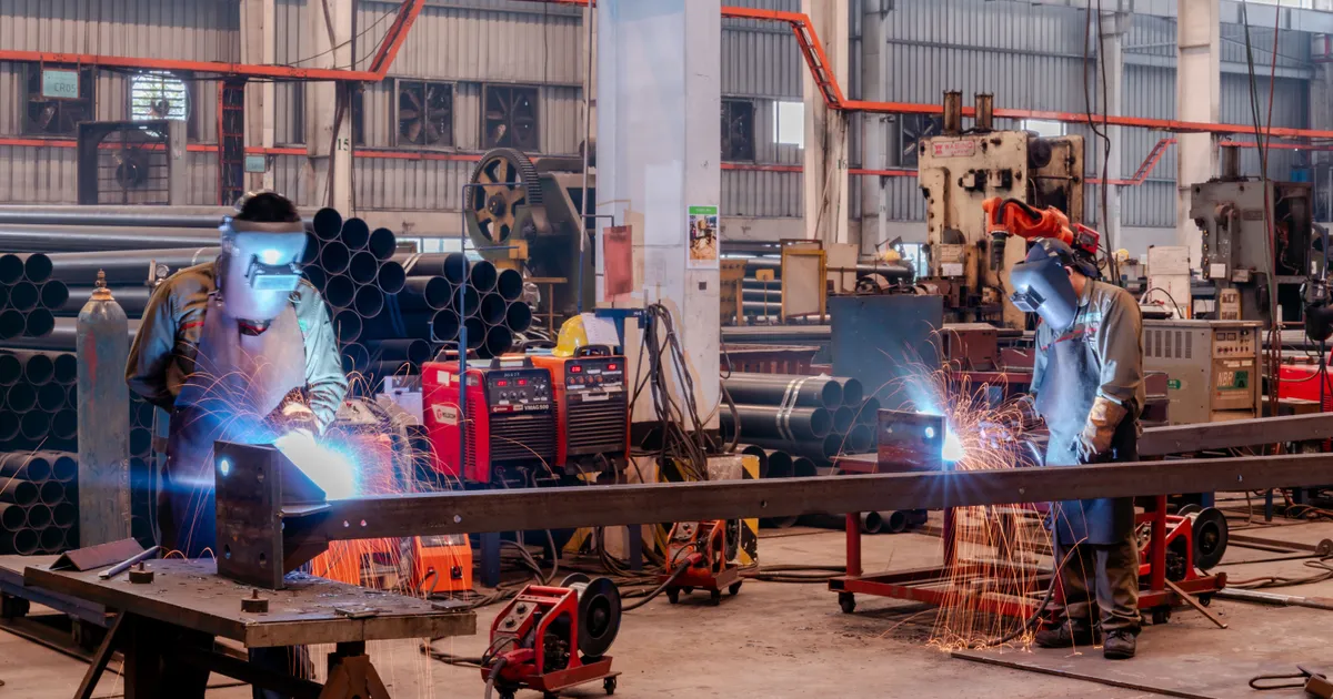 Dos soldadores con careta trabajan sobre vigas y perfiles de acero en una nave industrial, entre chispas, tanques de gas y equipos de soldadura