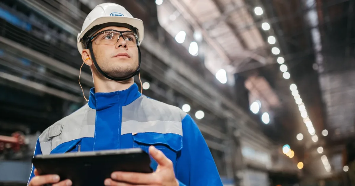 Supervisor de planta con casco blanco sostiene una tablet y observa la línea en el pasillo de una nave industrial