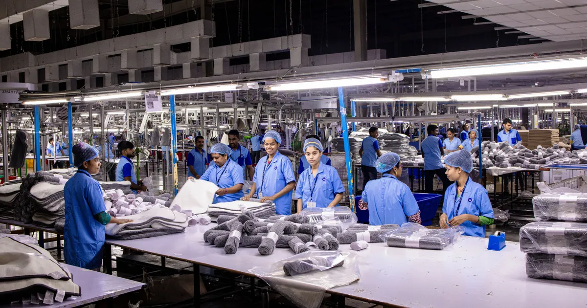 Nave de producción con decenas de operarias en bata azul y cofia empacando toallas dobladas en mesas largas bajo lámparas de techo
